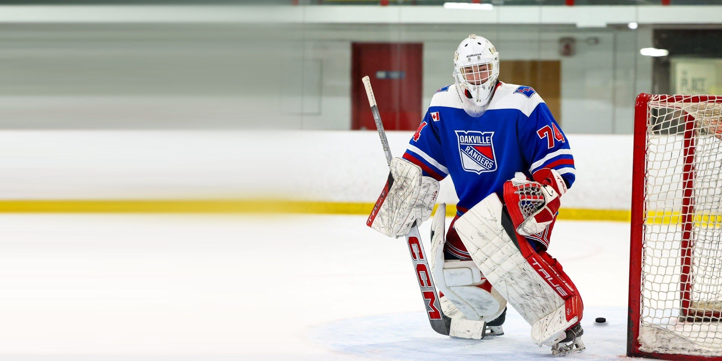 A goalie in a blue Oakville Rangers jersey sets in his stance beside the post, with a puck on the ice by the net.